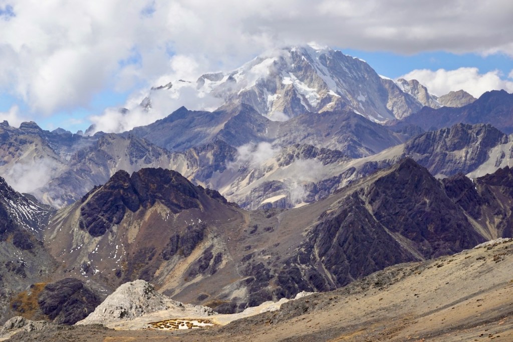 A panoramic view of rugged mountain peaks, showcasing a snow-capped summit surrounded by rocky slopes and clouds.