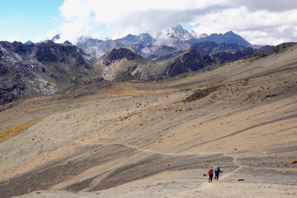 Two hikers trekking along a winding trail in a mountainous landscape, with rocky terrain and snow-capped peaks in the background under a partly cloudy sky.