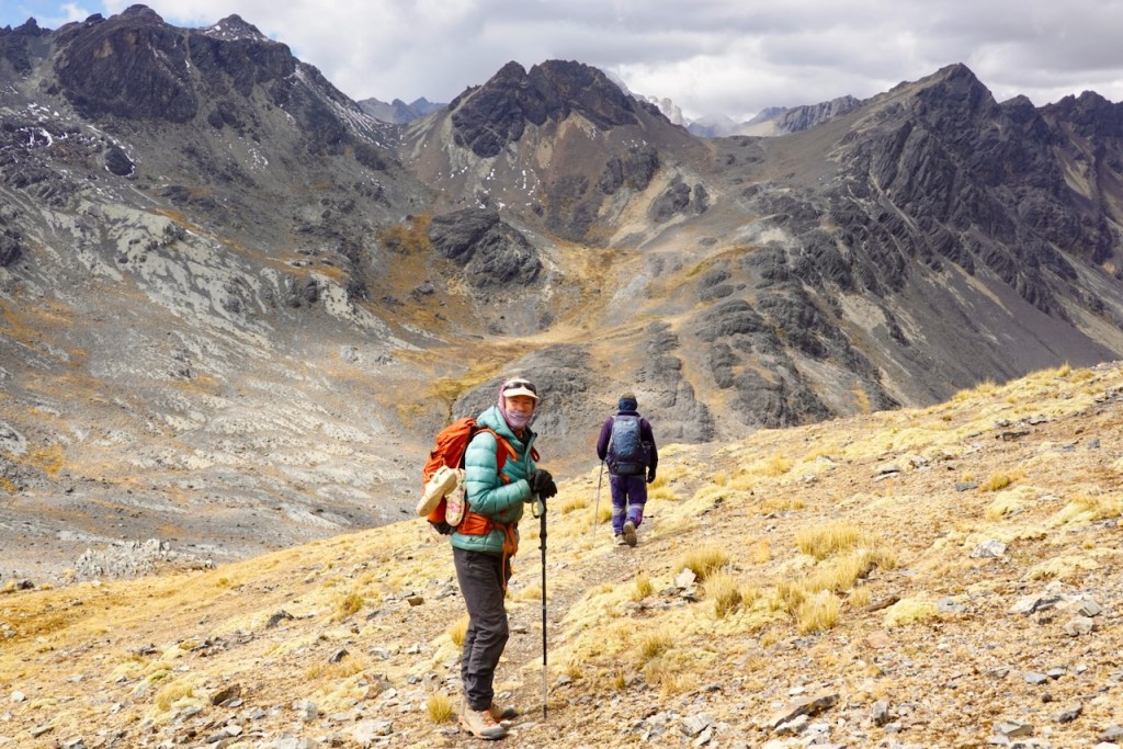 A hiker in a green jacket and orange backpack smiles at the camera while standing on a rocky hillside. In the background, another hiker in a blue jacket walks away towards a mountainous landscape with rocky terrain and scattered grass.