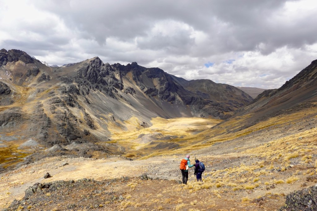 Two hikers walking through a rocky mountainous landscape with gray peaks and a valley below, set against a cloudy sky.