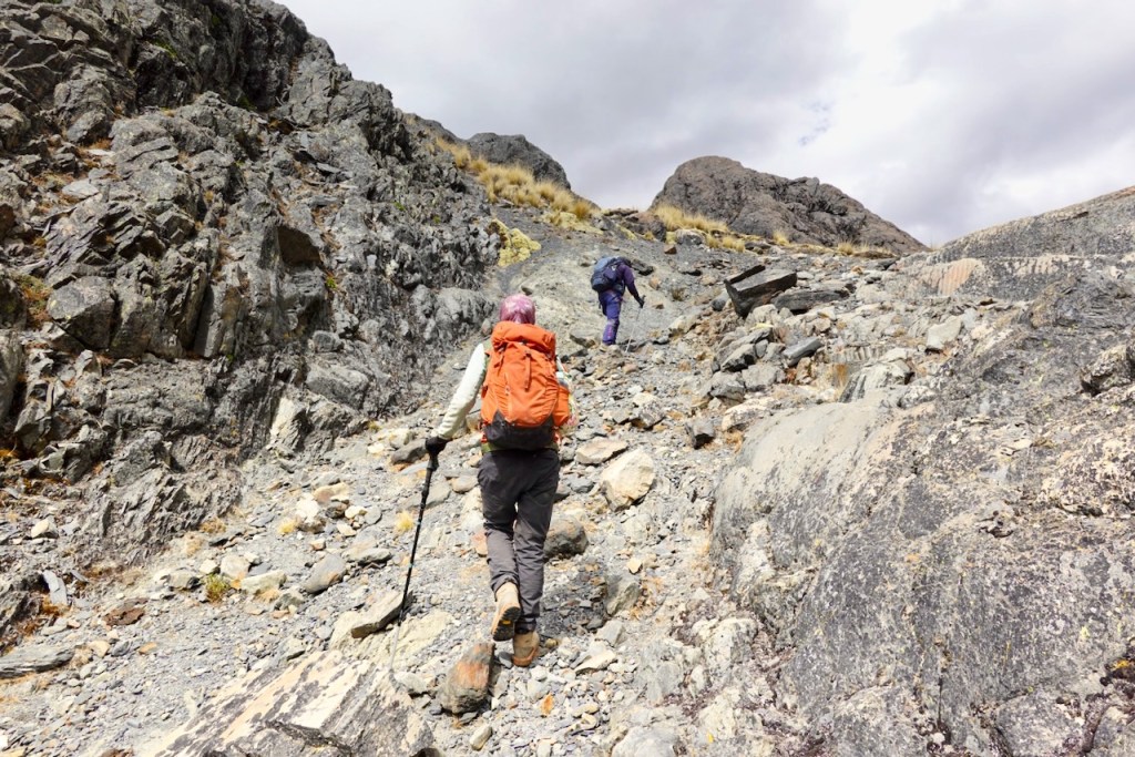 Two hikers climbing a rocky trail in a mountainous area, one with a red backpack and the other in purple gear.