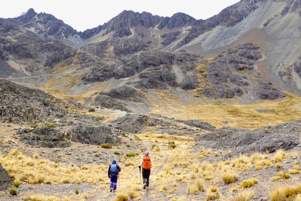 Two hikers walking on a rugged mountain trail surrounded by rocky terrain and sparse grassland, with towering peaks in the background.