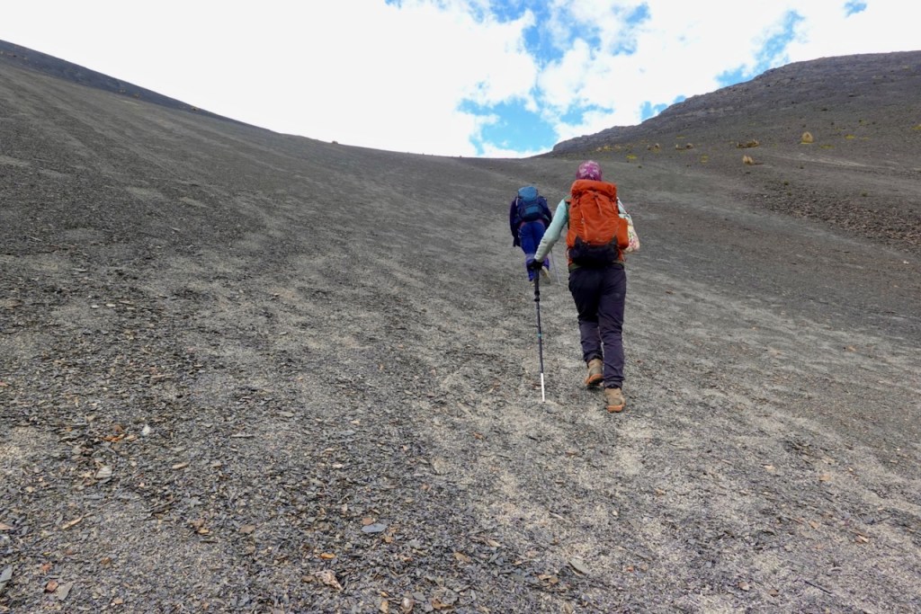 Two hikers ascending a steep, rocky slope under a partly cloudy sky.