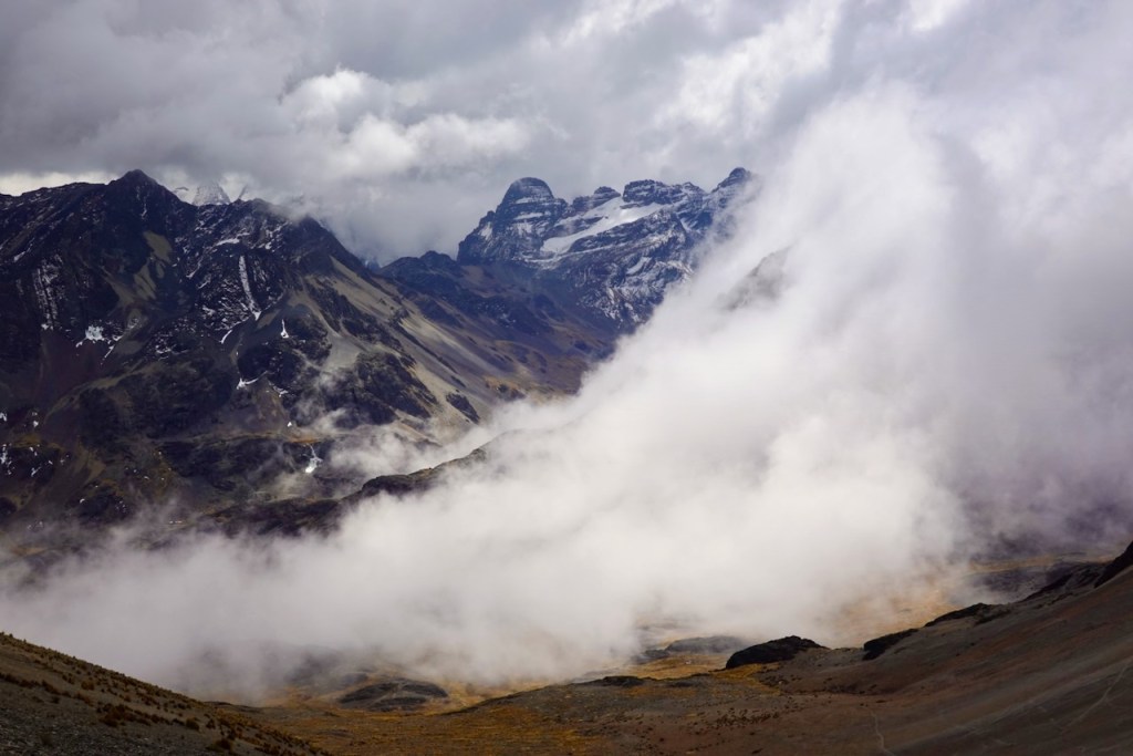 Dramatic mountain landscape with snow-capped peaks partially obscured by clouds and mist, showcasing rugged terrain and a valley below.