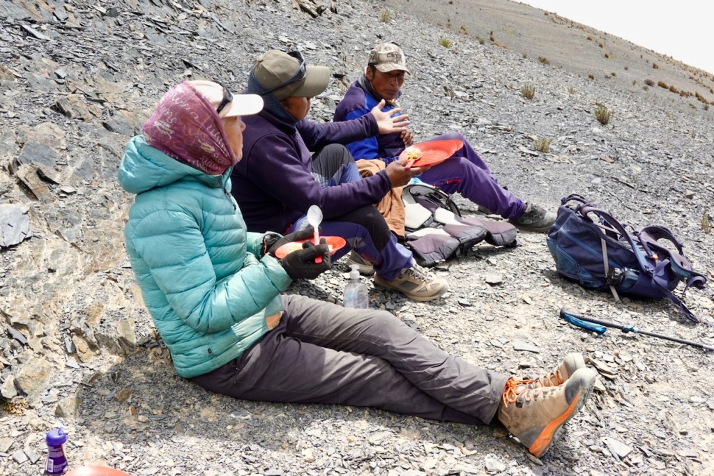 Three hikers taking a break on rocky terrain, enjoying a meal on red plates. They are dressed in outdoor attire, including jackets and hats.