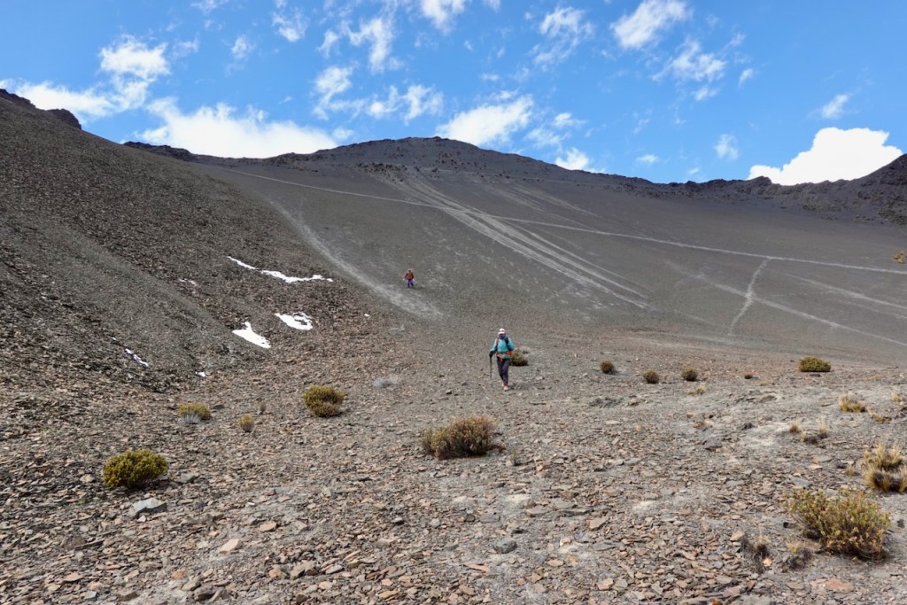 A hiker walking on a rocky slope with sparse vegetation, set against a backdrop of a steep hill and blue sky with clouds.