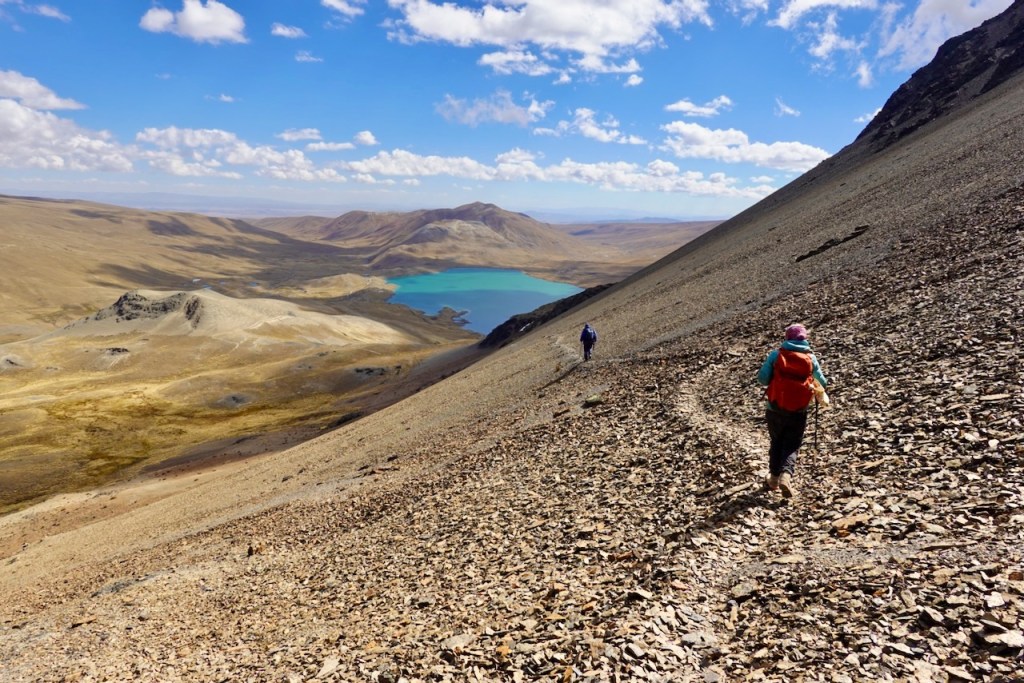 Two hikers descending a rocky slope with a turquoise lake and mountainous landscape in the background under a partly cloudy sky.