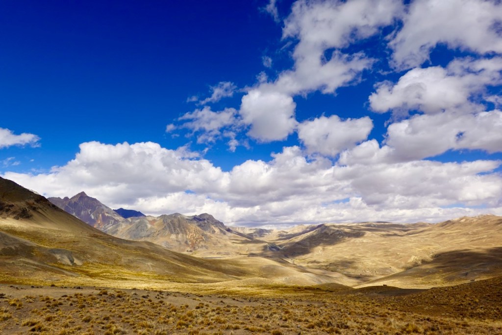 A panoramic view of a mountainous landscape under a bright blue sky with scattered white clouds, showcasing rugged terrain and dry grasslands.