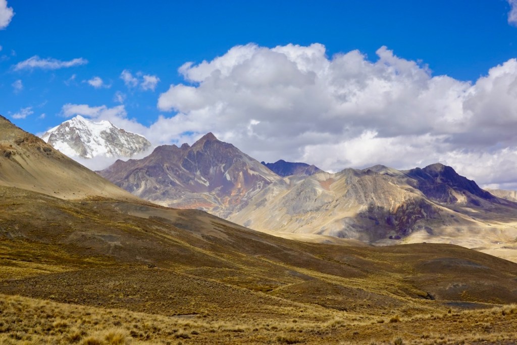 A panoramic view of rugged mountains with snow-capped peaks under a vibrant blue sky dotted with clouds. The foreground features arid terrain and the dramatic backdrop of various mountain ranges.