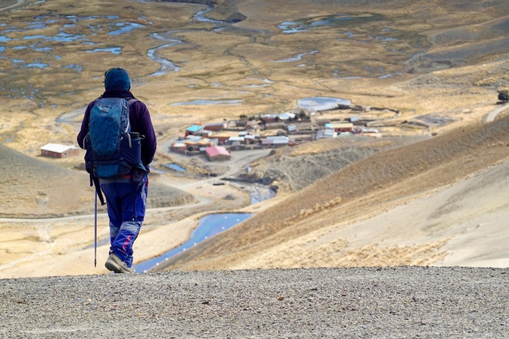 A hiker in outdoor gear walking on a rocky path, overlooking a remote village surrounded by hills and water bodies in a mountainous landscape.