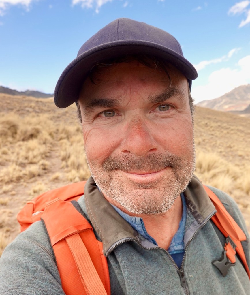 A smiling man with a beard and a hat standing in a dry, grassy landscape under a blue sky.