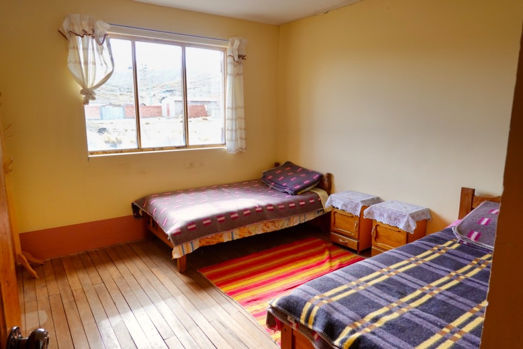 Interior of a simple bedroom featuring two beds with colorful blankets, wooden furniture, and a large window with curtains allowing natural light.