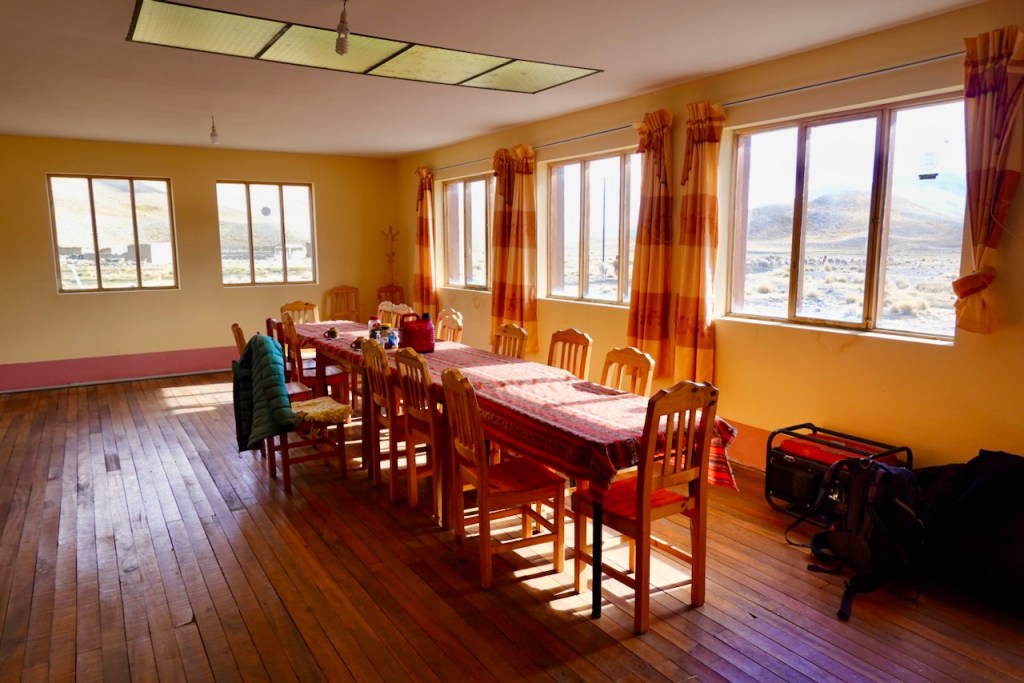 A spacious dining room with a long wooden table surrounded by chairs, set against a backdrop of large windows that let in natural light and offer views of a mountainous landscape. The floor is wooden, and the walls are painted a soft yellow.