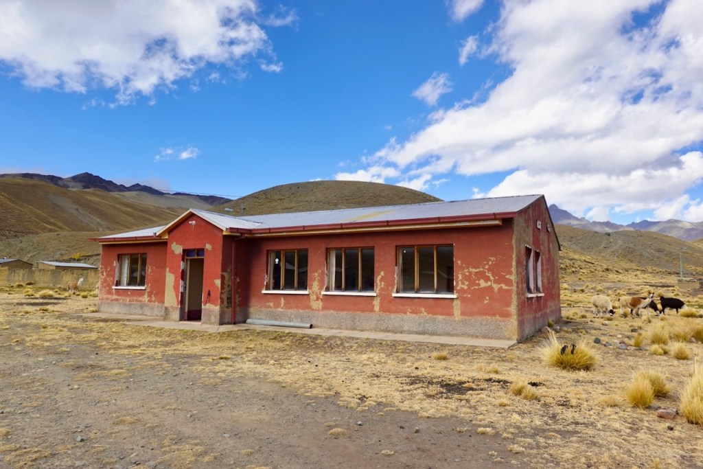 A weathered red building in a rural landscape, with mountains in the background and animals grazing nearby.