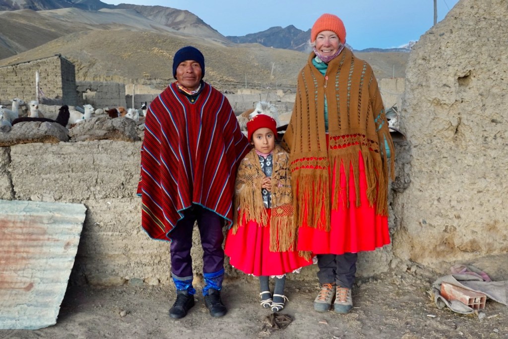 Three people standing together in traditional clothing: a man and a woman on either side, with a young girl in between. The woman wears a colorful shawl and a bright red skirt, while the man is dressed in a patterned poncho. They are in a rural setting with a stone wall and some animals in the background.