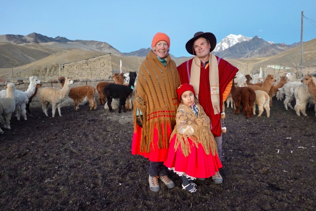 A family dressed in traditional clothing standing in a pasture surrounded by llamas and mountains in the background.