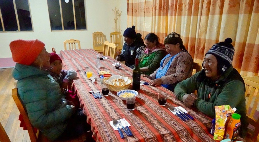 A group of six people, including adults and a child, sit around a long dining table filled with food and drinks in a cozy setting, featuring warm clothing and smiling faces.