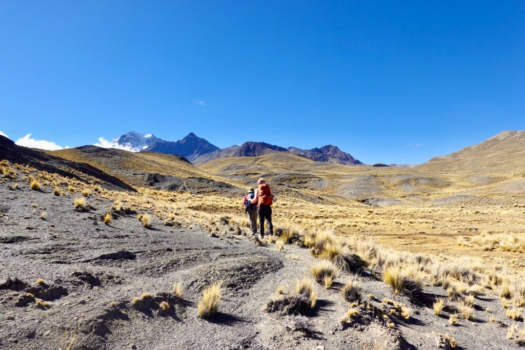 Two hikers walking on a rugged trail in a mountainous landscape under a clear blue sky.