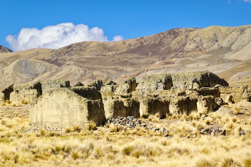 Ruins of ancient structures in a dry, mountainous landscape with blue sky and clouds.