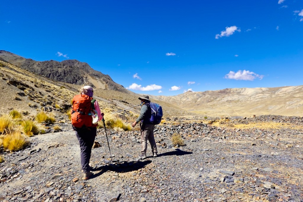 Two hikers with backpacks walking on a rocky trail in a mountainous desert landscape under a clear blue sky.