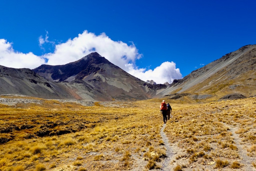 A hiker walking on a trail through golden grasslands, surrounded by rugged mountains under a clear blue sky with scattered clouds.
