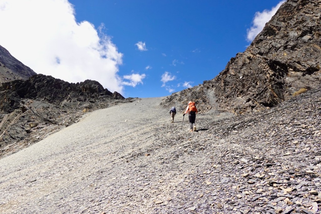 Two hikers trekking up a rocky slope under a blue sky with clouds.