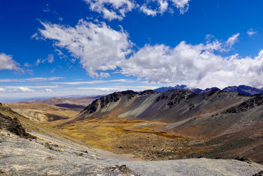 Panoramic view of a mountainous landscape featuring rugged terrain, sparse vegetation, and a blue sky with fluffy white clouds.