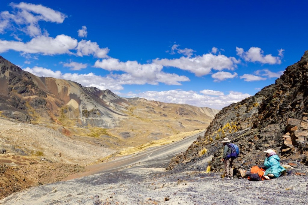 A scenic mountain landscape with hikers resting on rocky terrain, surrounded by colorful cliffs and a blue sky with fluffy clouds.