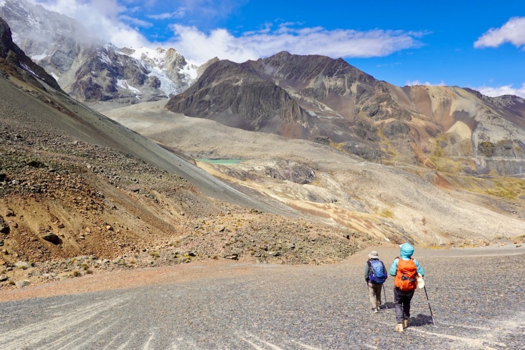 Two hikers walking on a rocky trail in a mountainous landscape with snow-capped peaks and blue sky.