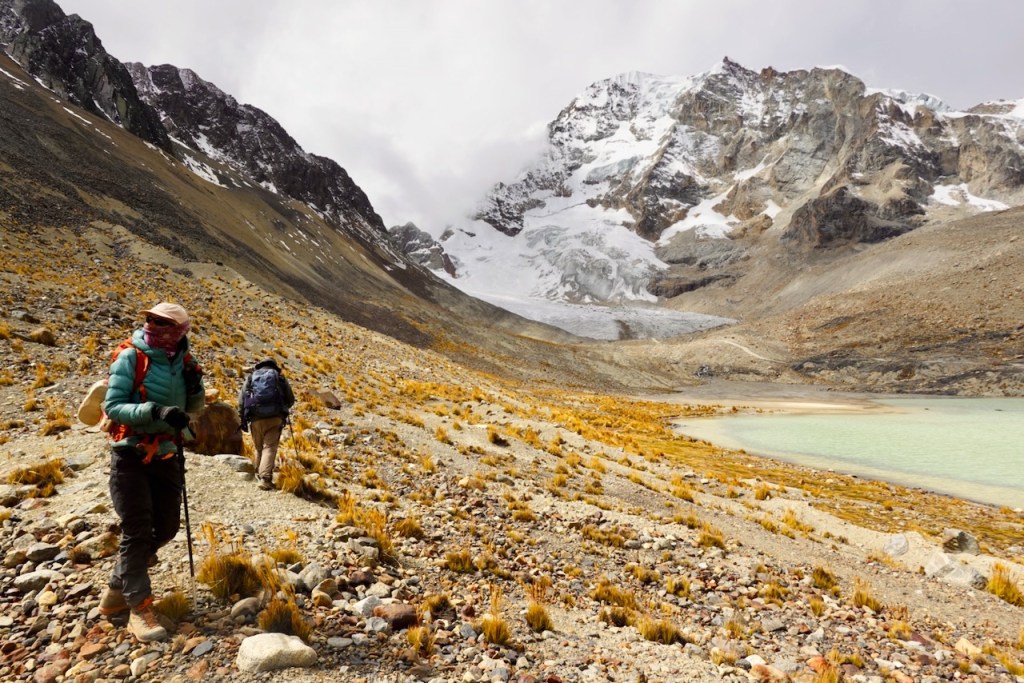 Two hikers trekking along a rocky trail in a mountainous landscape, with snowy peaks in the background and a glacial lake nearby.