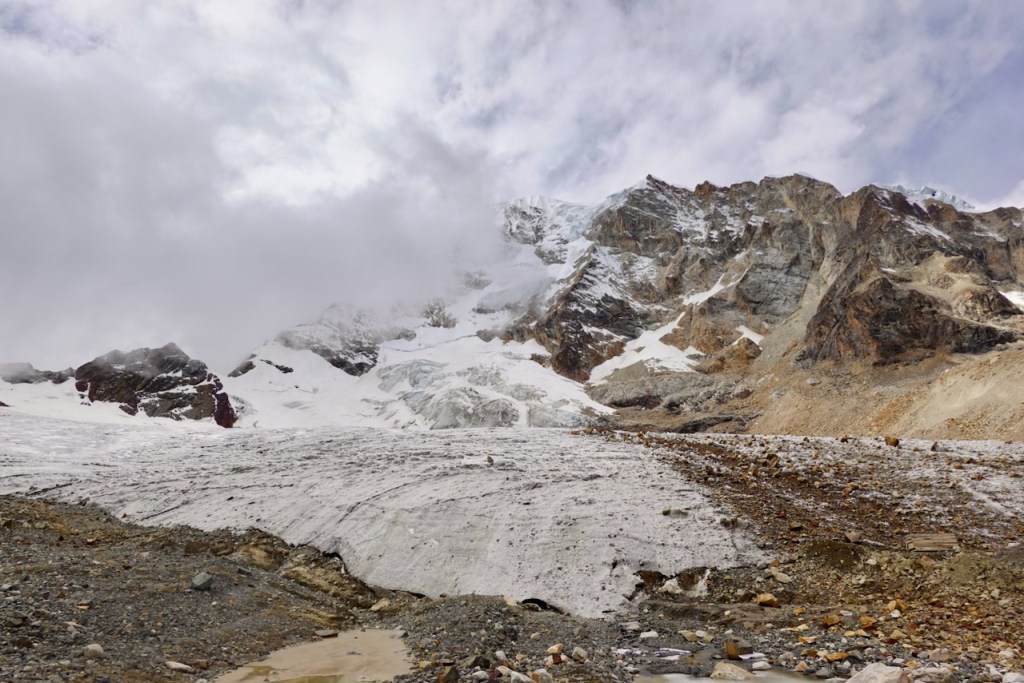 A mountainous landscape featuring a glacier in the foreground, with rocky terrain and clouds partially obscuring the peaks in the background.