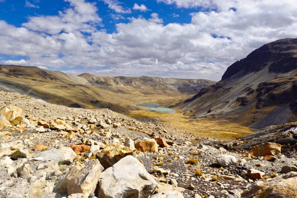 A panoramic view of a rocky mountainous landscape with a lake in the center, surrounded by rolling hills and a blue sky with scattered clouds.
