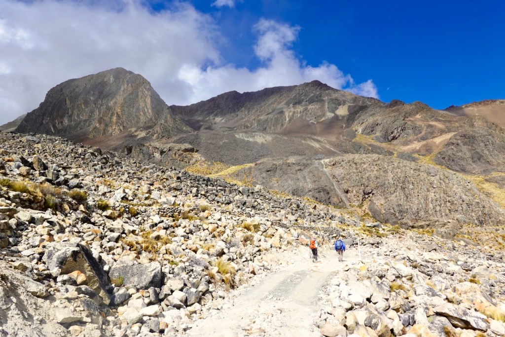 Two hikers walking on a rocky trail surrounded by mountains and a blue sky with clouds.