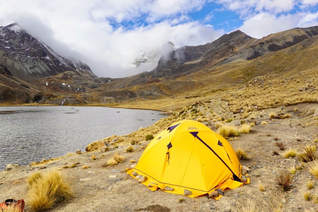 A bright yellow camping tent set up near a serene lake, surrounded by rugged mountains and grassland under a partly cloudy sky.