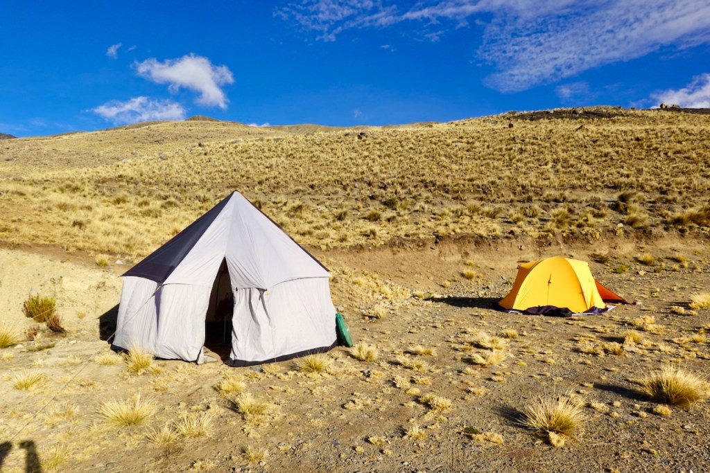 A large white tent and a smaller orange tent set in a dry, grassy landscape with blue skies and scattered clouds.