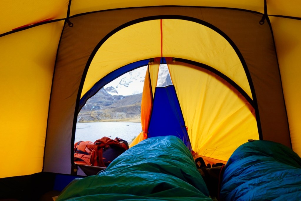 View from inside a colorful camping tent showing two sleeping bags and an outdoor landscape with mountains in the background.