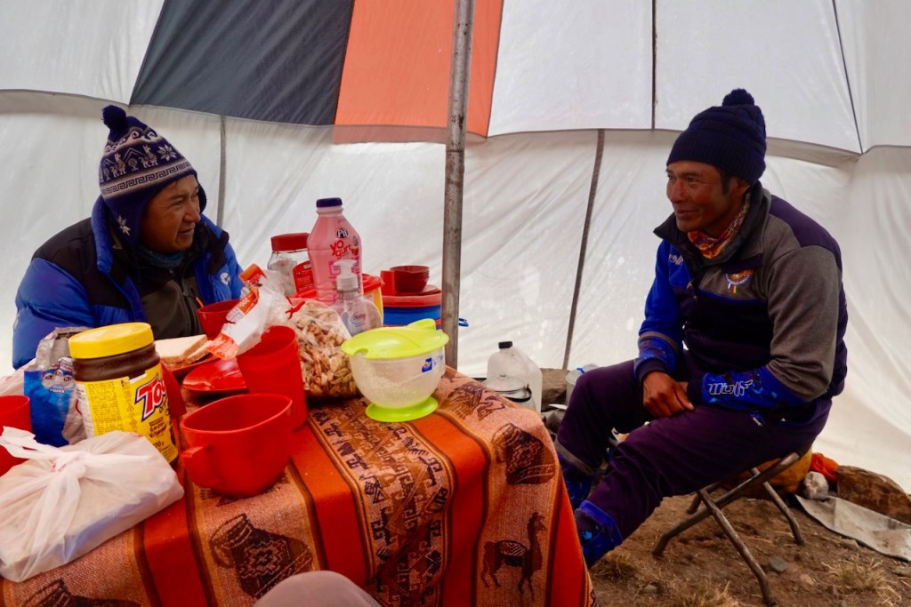 Two men sitting inside a tent, engaging in conversation. A table is filled with various food items and drinks, including red cups and containers.