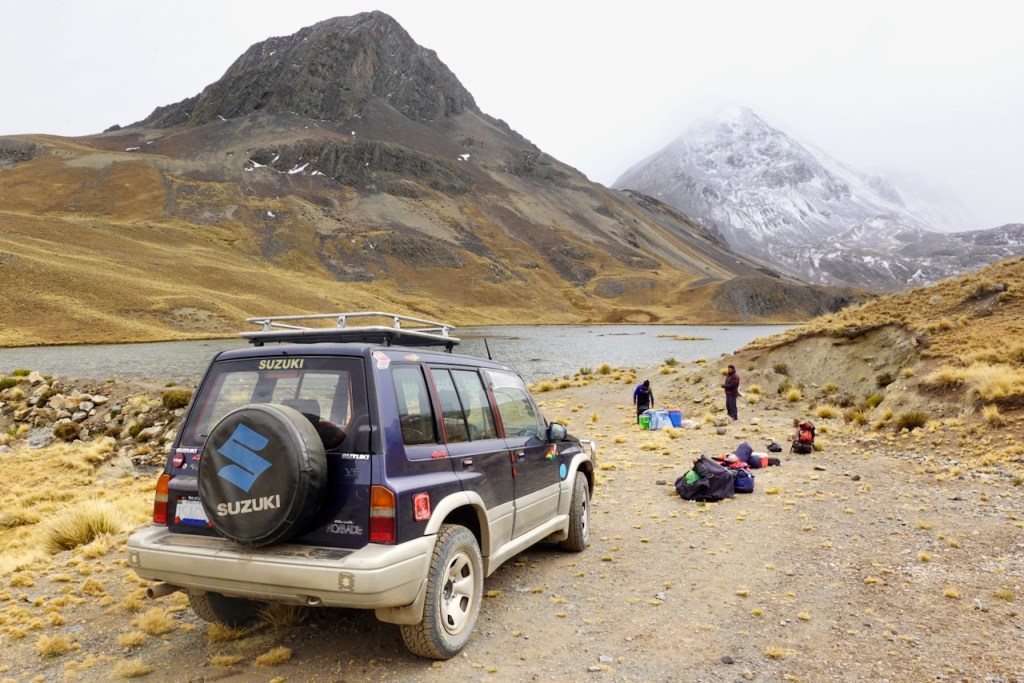 A Suzuki SUV parked near a lake in a mountainous landscape, with people setting up camping gear in the background.