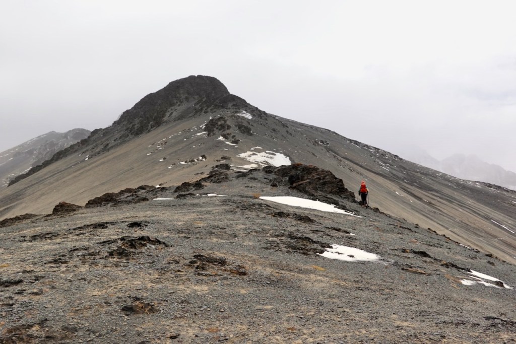 A hiker wearing an orange jacket climbing a rocky slope leading to a mountain peak under a cloudy sky.