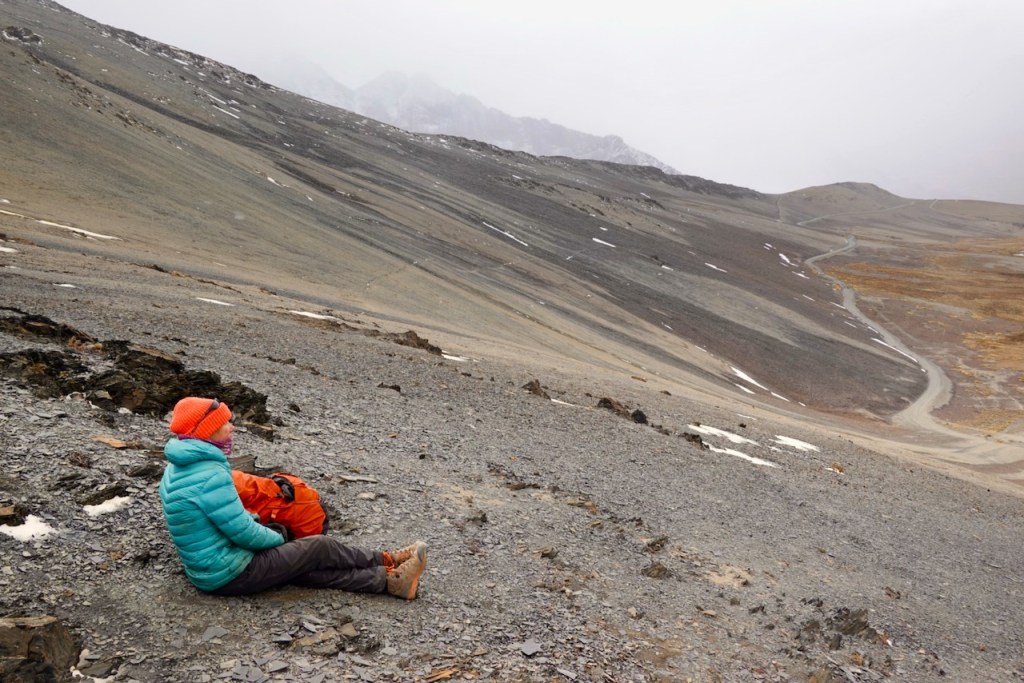 A person in a vibrant blue and orange outfit sits on a rocky slope, gazing at a distant winding road amidst a mountainous landscape shrouded in mist.