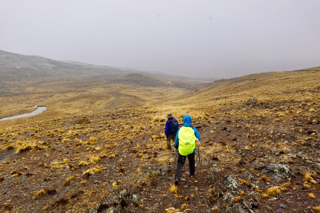 Two hikers trekking through a rocky, grassy landscape under a cloudy sky, with a winding river visible in the background.