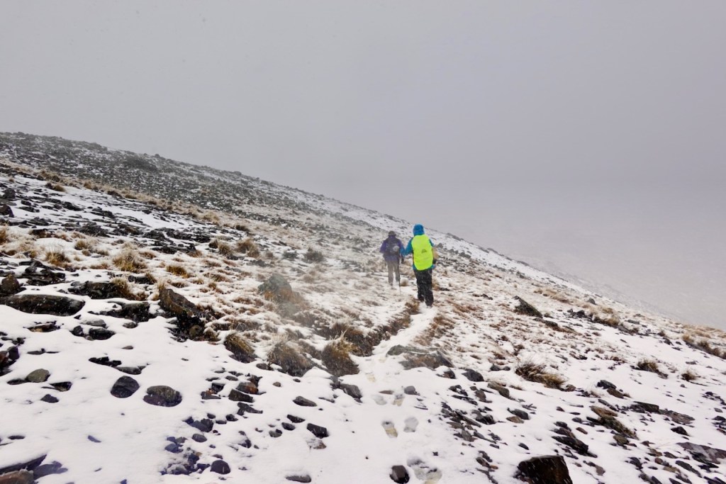 Two hikers trek up a snowy, rocky slope in low visibility conditions with a gray sky.