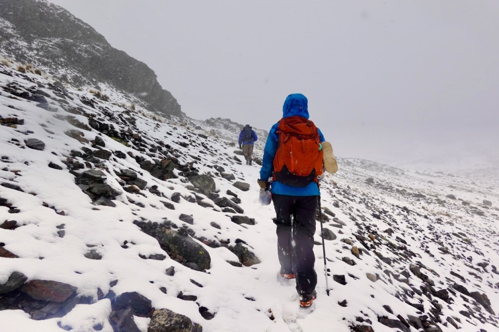 Two hikers trekking up a rocky, snow-covered slope in a mountainous area during winter weather.