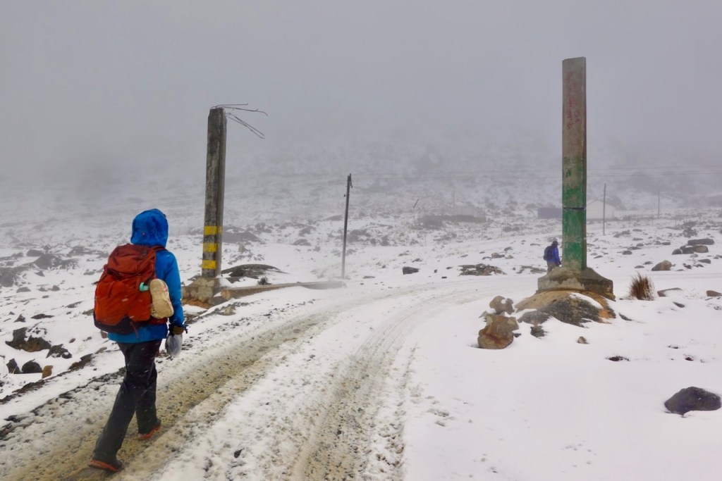 A hiker wearing a blue jacket and a brown backpack walks on a snowy road surrounded by fog and rocky terrain, with utility poles visible in the background.