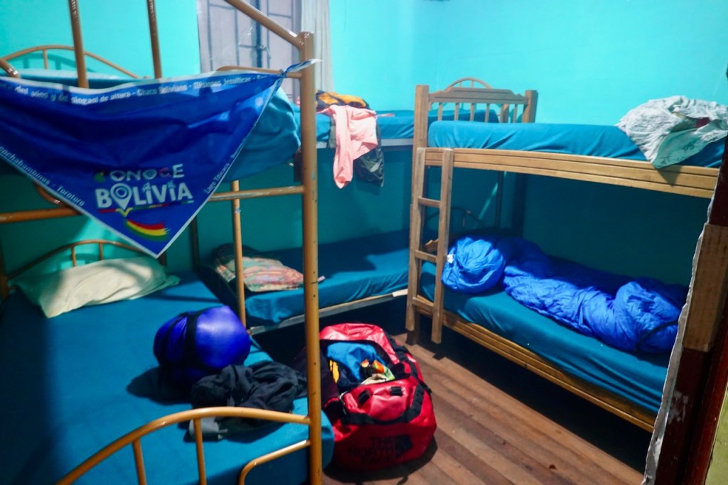 A shared dormitory room with two sets of wooden bunk beds, featuring blue mattresses and a brightly colored wall. A blue bandana promoting Bolivia is hanging from the upper bunk, while various personal items like sleeping bags and bags are scattered around the room.