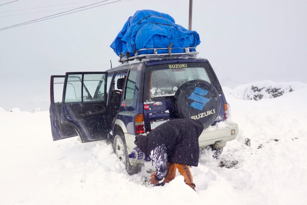 A Suzuki vehicle with a blue tarp on the roof is partially covered in snow, parked on a snowy landscape. A person in winter clothing is digging in the snow near the back of the vehicle, while the doors are open.