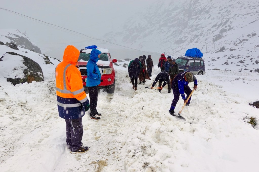 A group of people clearing snow from a road during a snowstorm, with vehicles parked in the background.