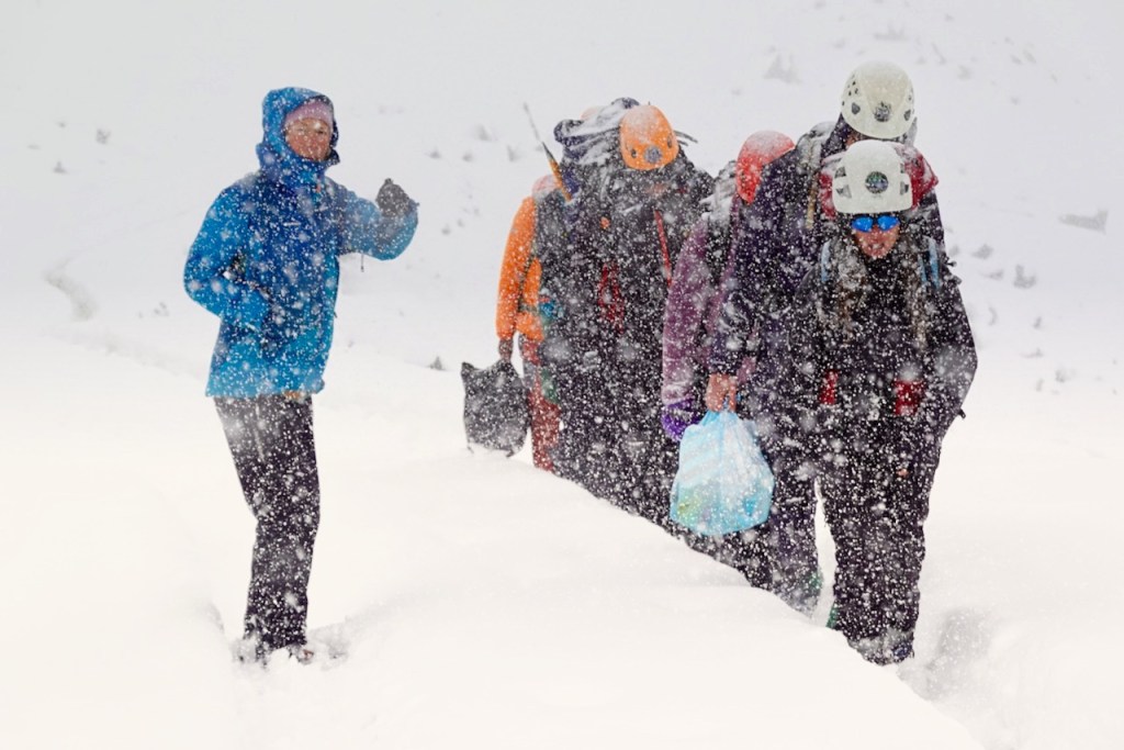 A group of climbers trekking through heavy snowfall on a glacier, with one person in a blue jacket leading the way.